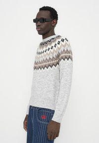 Ανοιχτό γκρι πλεκτό πουλόβερ με γεωμετρικό μοτίβο σε καφέ, μαύρο και κρεμ κατά μήκος του μανσέτου· ριμπ μανσέτες· σετ με σκούρο τζιν με λεπτές ρίγες.