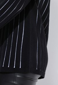 Schwarzer Strickpullover mit vertikalen weißen Streifen, gerippter Textur und lockerer Passform. Der Saum ist gerade und hinten etwas länger.