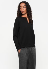 Zwarte pullover met een subtiele sluiting, lange mouwen en een relaxte pasvorm, gecombineerd met gestructureerde grijze high-waisted broeken.