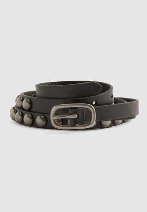 Ceinture en cuir noir avec des clous métalliques ronds et une boucle métallique rectangulaire, enroulée sur un fond uni gris clair.