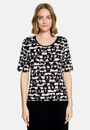 Femme aux cheveux blonds mi-longs portant une chemise rayée noire et blanche avec des taches léopard noires et un pantalon noir, debout sur fond blanc.