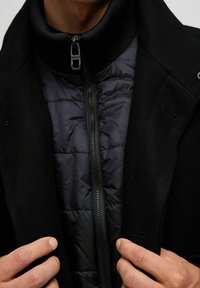 Svart ullkappa över en puffjacka. Kappan har en hög krage med dragkedja, medan puffjackan har en kviltad textur.