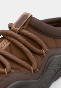 Zapatilla marrón con diseño acolchado. Presenta cordones prominentes, una suela texturizada y una base de goma. Talón reforzado con etiqueta de la marca.