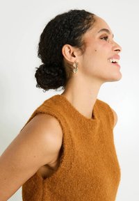 Femme aux cheveux bouclés en chignon bas, portant un haut sans manches en peluche brun et des boucles d'oreilles en or ornées, souriante et regardant vers la droite.