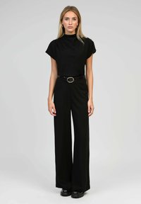 Combinaison noire avec un col haut, des manches courtes et une coupe ample. Accentuée par une ceinture noire avec une boucle ovale dorée. Texture lisse.