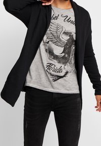 Musta neulejakku, jossa avoin edusta, yhdistettynä vaaleanharmaaseen t-paitaan, jossa on musta graafinen kuvio ja teksti. Mustat farkut, joissa on kevyt kulutus.