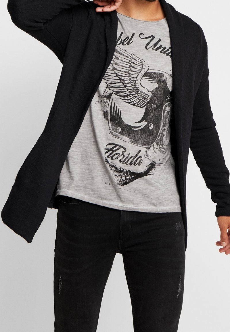 Musta neulejakku, jossa avoin edusta, yhdistettynä vaaleanharmaaseen t-paitaan, jossa on musta graafinen kuvio ja teksti. Mustat farkut, joissa on kevyt kulutus.