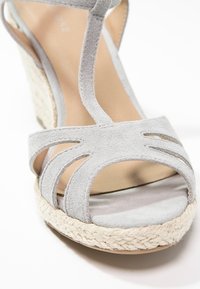 Sandale compensée en daim gris avec découpes ouvertes, une doublure intérieure douce, et une semelle en jute tressé et caoutchouc pour plus de texture et de stabilité.