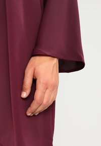 Burgundytyg med en slät, lätt glänsande yta. Bilden visar en hand som vilar på en bred, uppvikbar ärm.