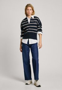 Jūras spēku svītrains adīts džemperis ar V veida kakla izgriezumu, zem tā balta apkakles krekla, apvienots ar tumši zilām platām biksēm un baltām kedām.