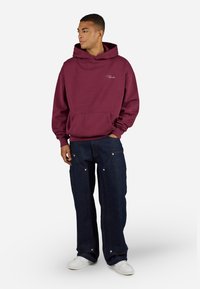 Maroon hoodie with kangaroo pocket, relaxed fit, ribbed cuffs, paired with dark blue denim pants featuring exposed seams and metal accents.