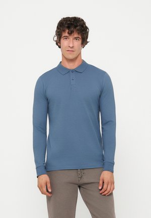 Homme aux cheveux bouclés portant un polo bleu à manches longues et un pantalon marron clair, debout devant un fond blanc uni.