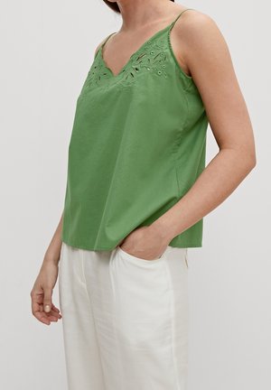 Femme portant un haut vert sans manches avec un col brodé et un pantalon blanc, une main dans la poche, sur un fond uni.