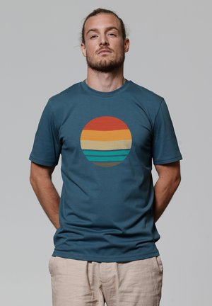 Teal katoenen T-shirt met een ronde grafische print met horizontale strepen in rood, oranje, geel, teal en bruin. Korte mouwen en ronde halslijn.