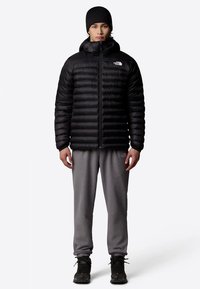 Jaunuolis, dėvintis juodą pūkinę striukę, juodą kepuraitę, pilkas sportines kelnes ir juodus žygių batus, stovintis priešais paprastą šviesų foną.