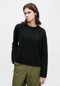 Νεαρή γυναίκα με ίσια καστανά μαλλιά, που φοράει μια μαύρη μπλούζα με μακριά μανίκια και ελαιοπράσινα cargo παντελόνια, στέκεται μπροστά σε λευκό φόντο.