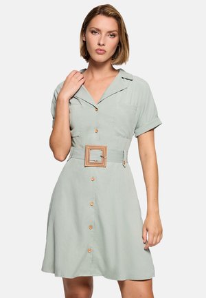 Mujer con un vestido verde claro abotonado, mangas cortas remangadas y un cinturón ancho tejido con una hebilla cuadrada.