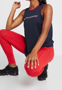 Marinblå atletisk linne med röda detaljer, prydd med en logotyp; kombineras med kroppsnära röda leggings; svarta sneakers fulländar outfiten.