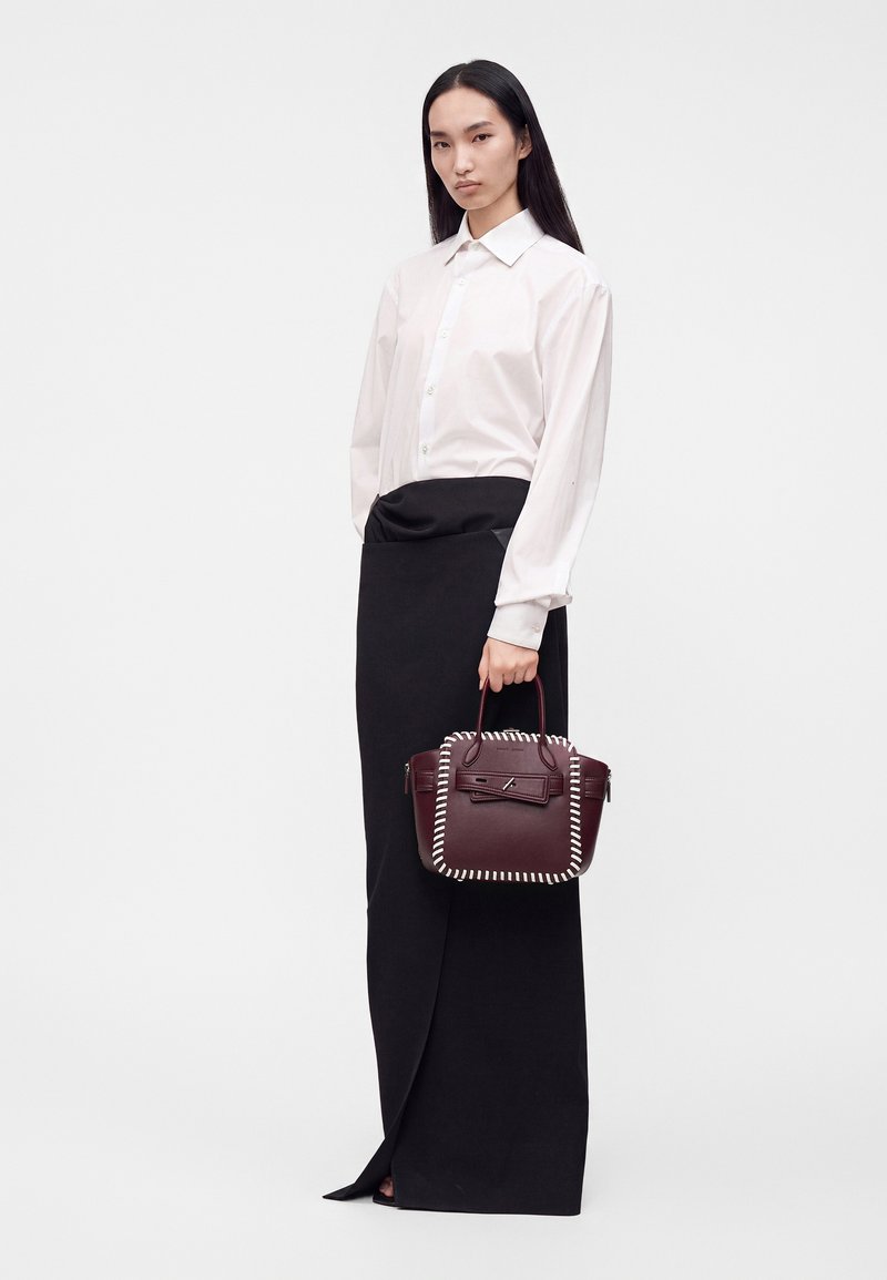 Femme aux longs cheveux noirs portant une chemise blanche boutonnée et une jupe noire, tenant un sac à main rouge foncé avec des détails de couture blancs.
