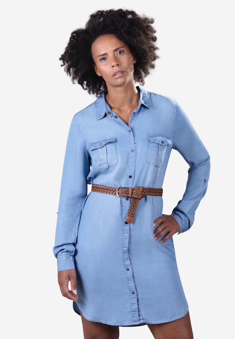 Vrouw met krullend haar die een lichtblauwe denim jurk met korte mouwen en een bruine gevlochten riem draagt, poserend met één hand op haar heup tegen een witte achtergrond.