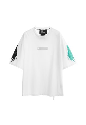 Joven con camiseta blanca de gran tamaño, pantalones deportivos negros con diseños en color teal y botas blancas con cordones, de pie frente a un fondo blanco.