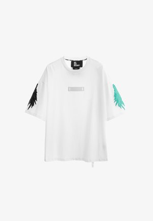 Joven con camiseta blanca de gran tamaño, pantalones deportivos negros con diseños en color teal y botas blancas con cordones, de pie frente a un fondo blanco.
