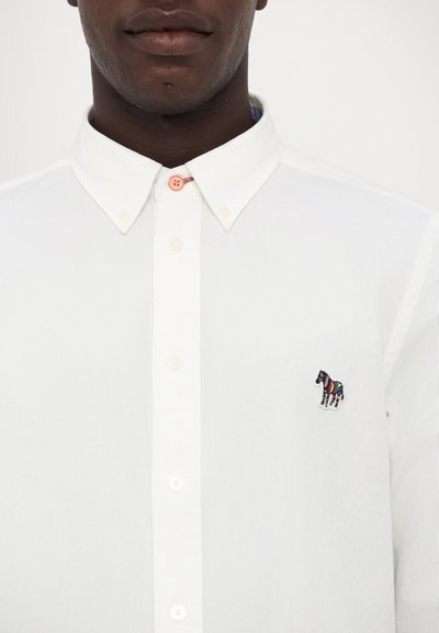 Vit skjorta med stiv krage, med en liten, flerfärgad broderad zebra-design på vänster bröst.