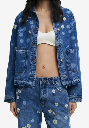 Blaue Jeansjacke und Jeans mit weißen Gänseblümchen-Prints, getragen über einem weißen, bauchfreien Oberteil mit Knopfleiste vorne.