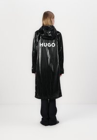 Melnā, spīdīgā garā mētelī ar kapuci un balto "HUGO" logo aizmugurē. Ar teksturētu virsmu, taisnu griezumu un elastīgiem apmaliem.
