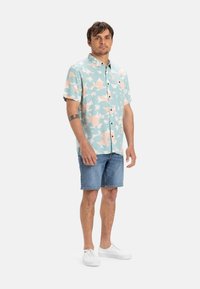 Camicetta a maniche corte di colore azzurro chiaro con motivo floreale rosa, con chiusura a bottoni sul davanti, abbinata a pantaloni corti in denim e sneakers bianche.