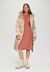 Beige gefütterter Mantel mit Kapuze, geripptem rotem Kleid und cremefarbenen Ankle Boots. Merkmale eines mittellangen Designs mit glatter Textur und seitlichen Taschen.