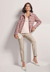 Roze tweedjasje met multikleurige accenten, gecombineerd met een lichtroze top en beige slim fit broek. Accessoires omvatten een clutch en witte hakken.