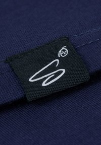 Marineblauwe stof met een gladde textuur, voorzien van een zwart label met witte stiksels en een gestileerd logo.