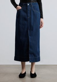 Τζιν φούστα σε σκούρο μπλε με ίσια γραμμή, εμπρόσθια σχισμή και εμφανείς ραφές. Διαθέτει εμπρόσθιες τσέπες και κλείσιμο με κουμπιά.