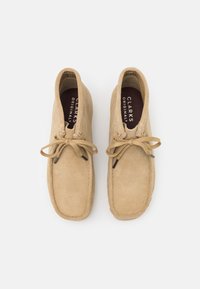 Barna velúr cipők lekerekített orral, fűzős elülső résszel és látható varrással. A talpbetéten "CLARKS ORIGINALS" márkázás található.