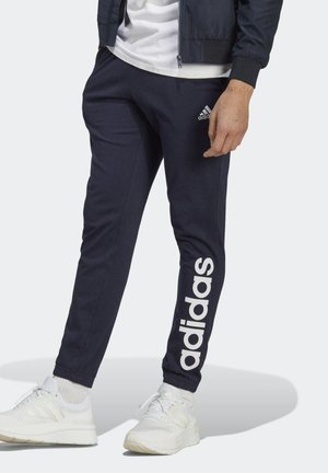Mand iført marineblå Adidas træningsbukser med hvidt logo og hvide sneakers, stående mod en ensartet lys baggrund.