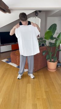 Una t-shirt bianca oversize tenuta da una persona, realizzata in tessuto con una consistenza liscia. La maglietta presenta maniche corte e un colletto arrotondato.