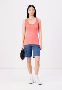 Top de tirantes de coral con ribetes de encaje, combinado con pantalones cortos de mezclilla hasta la rodilla. La modelo sostiene un bolso negro y lleva zapatillas altas blancas.