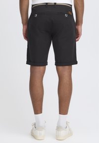Shorts noirs sur mesure avec ourlet roulé, poches arrière avec fermeture à bouton, et taille contrastante. Portés avec des chaussettes blanches et des baskets.