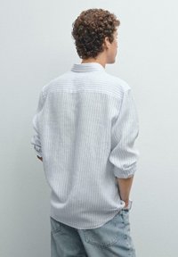Chemise rayée en gris clair et blanc, manches longues, coupe décontractée, avec des poignets retroussés et un col classique. Portée avec un short en denim clair.