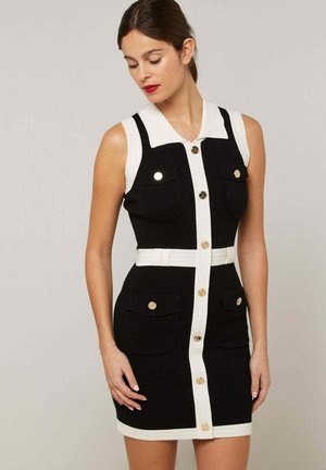 Vrouw draagt een mouwloze zwarte jurk met witte biezen, gouden knopen, een kraag, vier zakken en een bijpassende witte riem.