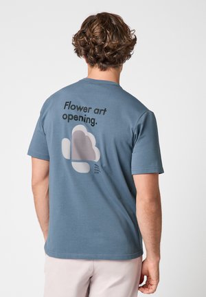 Uomo con capelli ricci castani indossa una maglietta blu con la scritta "Flower art opening" e un design floreale astratto sul retro, abbinata a pantaloni chiari.