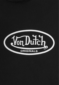 Svart tyg med en vit oval logotyp som säger "Von Dutch ORIGINALS" i stiliserad font med dekorativa accenter.