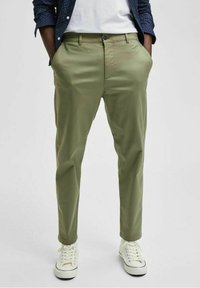 Green chinos with a smooth texture, featuring a standard fit, front pockets, and a button closure. Paired with white sneakers.