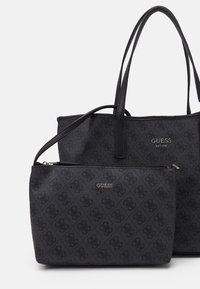 Deux sacs à main GUESS noirs avec un motif de logo subtil, un tote avec de longues poignées et un sac à bandoulière zippé plus petit.