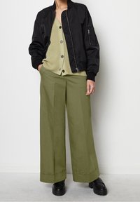 Chaqueta bomber negra, cárdigan verde acanalado con botones, pantalones anchos de pierna ancha color oliva claro y zapatos negros gruesos. Texturas simples y líneas limpias.