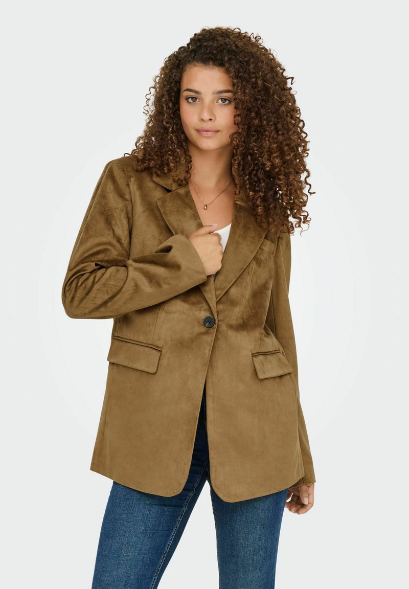 Bruine, zachte suède blazer met een enkele knoopsluiting, notch revers en twee voorkatoen. Draagt over een witte top en denim jeans.