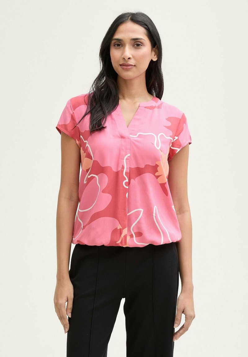Rosa Bluse mit abstraktem Blumenmuster, kurzen Ärmeln, V-Ausschnitt und gerafftem Saum; getragen mit schwarzen Hosen. Glattes Material, lockere Passform.