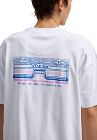 White cotton t-shirt featuring a blue graphic of a stadium and text reading "SERVING THE GAME FOR GENERATIONS" in blue and pink.