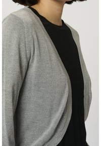 Cardigan argenté en tissu tricoté scintillant, avec une ouverture sur le devant, une coupe décontractée et une texture douce, porté sur une chemise noire.
